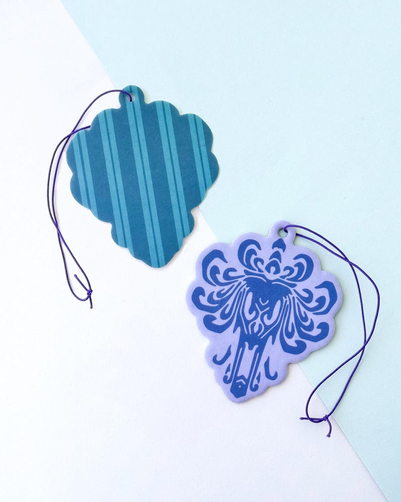 May include: Two car air fresheners, one with a blue and white striped pattern and the other with a purple background and a blue silhouette of a ghost.