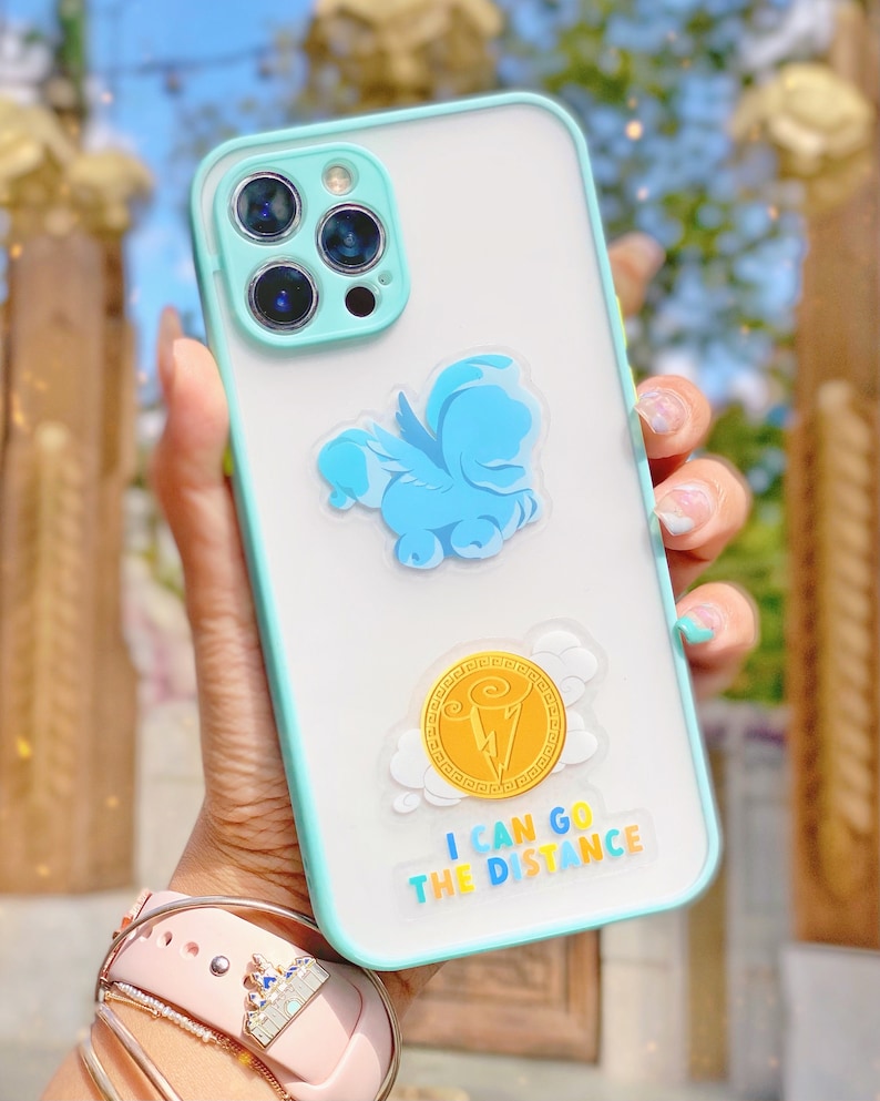 Puede incluir: Funda transparente para tel&eacute;fono con borde y detalles de c&aacute;mara en azul claro. La funda presenta un Pegaso de dibujos animados azul y un gr&aacute;fico de moneda de oro con el texto "I CAN GO THE DISTANCE".