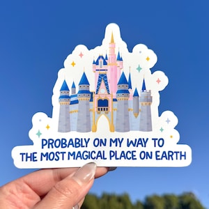 May include: A sticker featuring a cartoon illustration of a pink and blue castle with the text "PROBABLY ON MY WAY TO THE MOST MAGICAL PLACE ON EARTH" in white and blue. The sticker has a white border and is held against a blue sky.