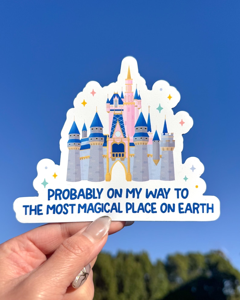 May include: A sticker featuring a cartoon illustration of a pink and blue castle with the text "PROBABLY ON MY WAY TO THE MOST MAGICAL PLACE ON EARTH" in white and blue. The sticker has a white border and is held against a blue sky.