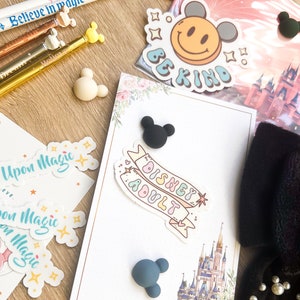 May include: A white card with a pink and green floral design. The card has a black and white sticker that says "Disney Adult" and a sticker with a yellow smiley face that says "Be Kind". There are also several small black and blue Mickey Mouse ears.