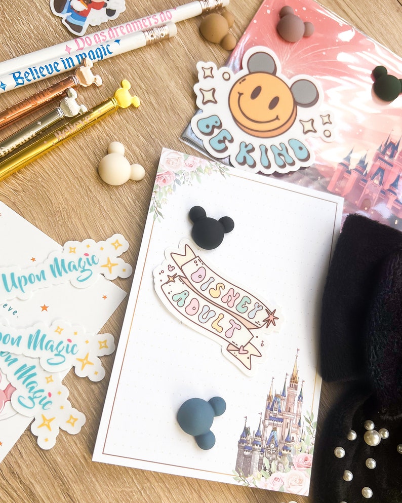 May include: A white card with a pink and green floral design. The card has a black and white sticker that says "Disney Adult" and a sticker with a yellow smiley face that says "Be Kind". There are also several small black and blue Mickey Mouse ears.