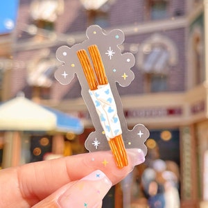 May include: A clear sticker with a cartoon illustration of two churros with a white frosting and blue sprinkles. The sticker has a sparkly background with white stars and blue and yellow glitter.