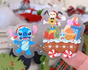 May include: A clear sticker featuring Stitch pulling a wagon filled with gifts and Pluto. Stitch is blue with a Santa hat and Christmas lights. Pluto wears a Santa hat and holds a bone. The wagon has candy wheels.