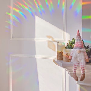 May include: A white wall with a rainbow prism effect. A white mantelpiece with two white candles and a pink and white gnome sitting on it.