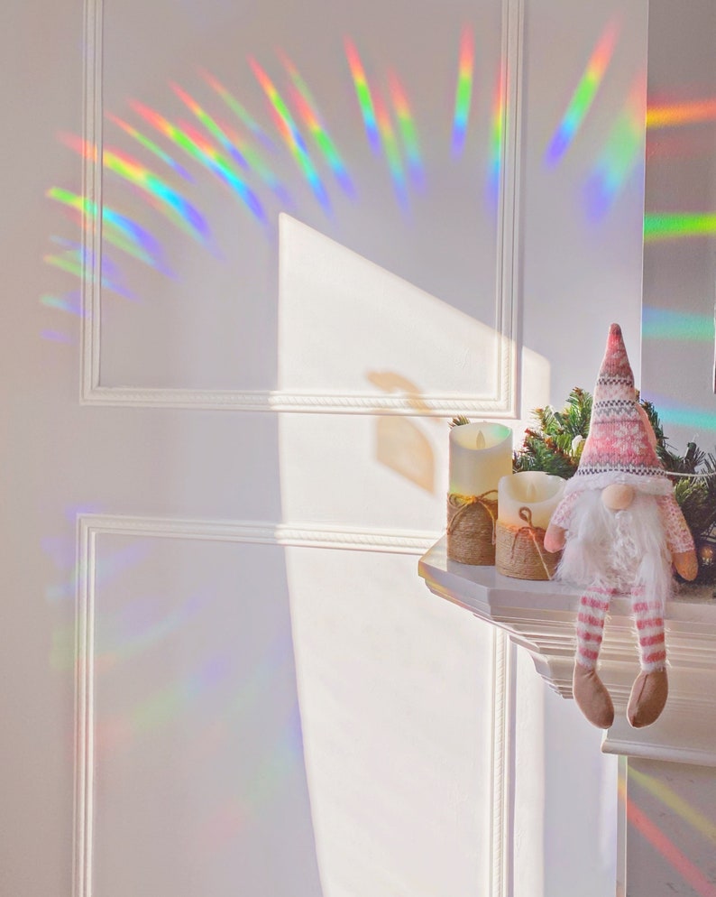 May include: A white wall with a rainbow prism effect. A white mantelpiece with two white candles and a pink and white gnome sitting on it.