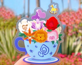 Puede incluir: Una taza de té azul con un platillo blanco y un arreglo floral en su interior. Las flores son amarillas, naranjas, rosas, blancas y moradas. Hay un pequeño caballo de balancín amarillo sobre las flores.