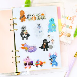 May include: A sheet of clear stickers featuring various Disney and Star Wars characters. The stickers include Chewbacca, R2-D2, BB-8, a droid, a blue monster, a Mandalorian with Grogu, a white furry creature, Darth Vader with a Stormtrooper, a purple dragon, a purple monster with a hat, a red wagon with colorful balls, and Stitch holding Mickey Mouse.