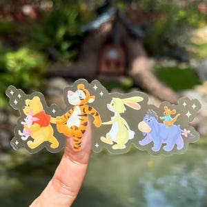 May include: A clear sticker featuring Winnie the Pooh, Piglet, Tigger, Rabbit, Eeyore, and Roo. The characters are in a line, holding hands. The sticker has a decorative border with small star shapes.