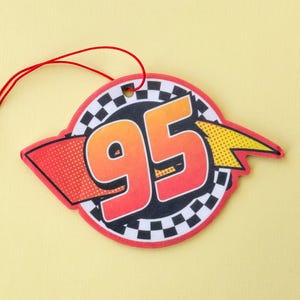 May include: Red, yellow, and black car air freshener with the number 95 on it. The air freshener is shaped like a circle with a checkered border.