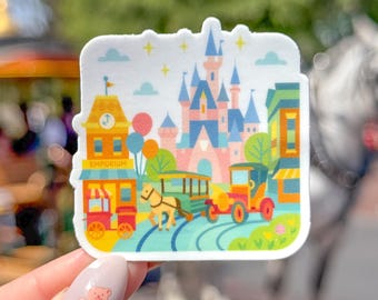 May include: A colorful sticker featuring a whimsical theme park scene. The design includes a pink and blue castle, a horse-drawn carriage, and a train. The sticker has a white border and is held up by a person.
