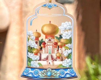 May include: A clear glass dome sticker with a detailed illustration of a golden palace with white flowers surrounding it. The palace has a blue and gold design with a blue gem at the base.