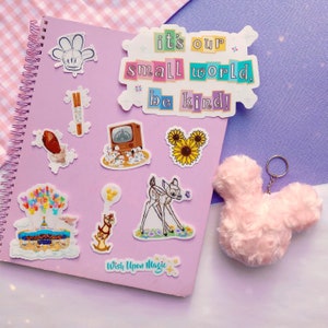 May include: A collection of stickers and a pink fluffy keychain shaped like a mouse head on a purple spiral notebook. The stickers feature various designs, including a deer, sunflowers, and text that reads "it's our small world, be kind!"