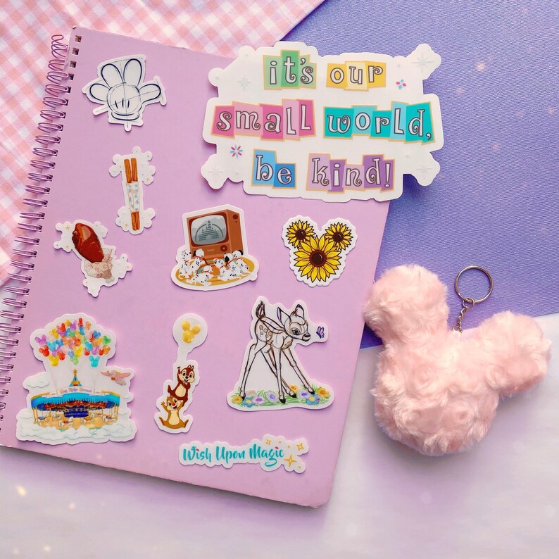 May include: A collection of stickers and a pink fluffy keychain shaped like a mouse head on a purple spiral notebook. The stickers feature various designs, including a deer, sunflowers, and text that reads "it's our small world, be kind!"