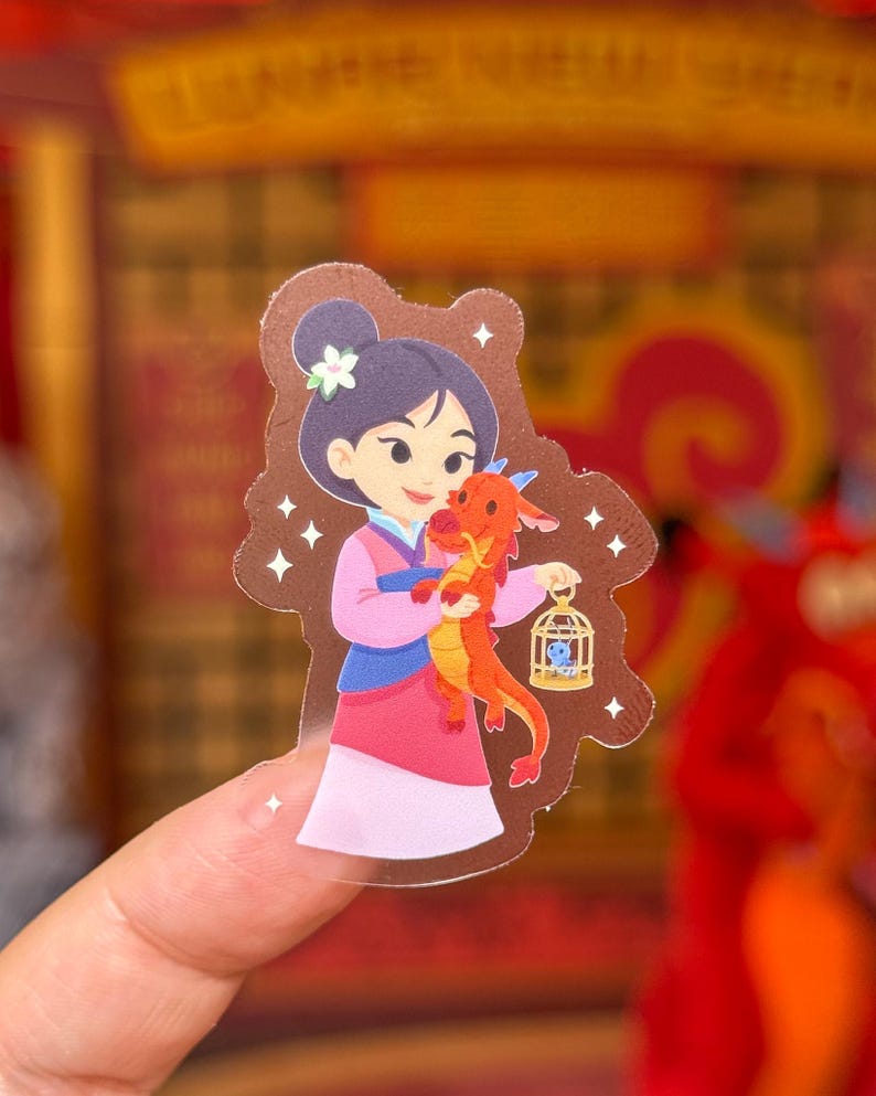 May include: A clear sticker featuring a cartoon illustration of a character in a pink and blue outfit holding a red dragon. The dragon holds a small cage. The character has dark hair and a flower. White star accents surround the image.