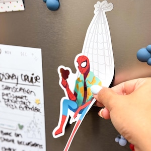May include: A colorful Spider-Man magnet, holding a treat, is depicted sitting on a white web. The superhero is wearing a Hawaiian shirt, blue pants, and red boots. The magnet is being held up against a metallic surface.