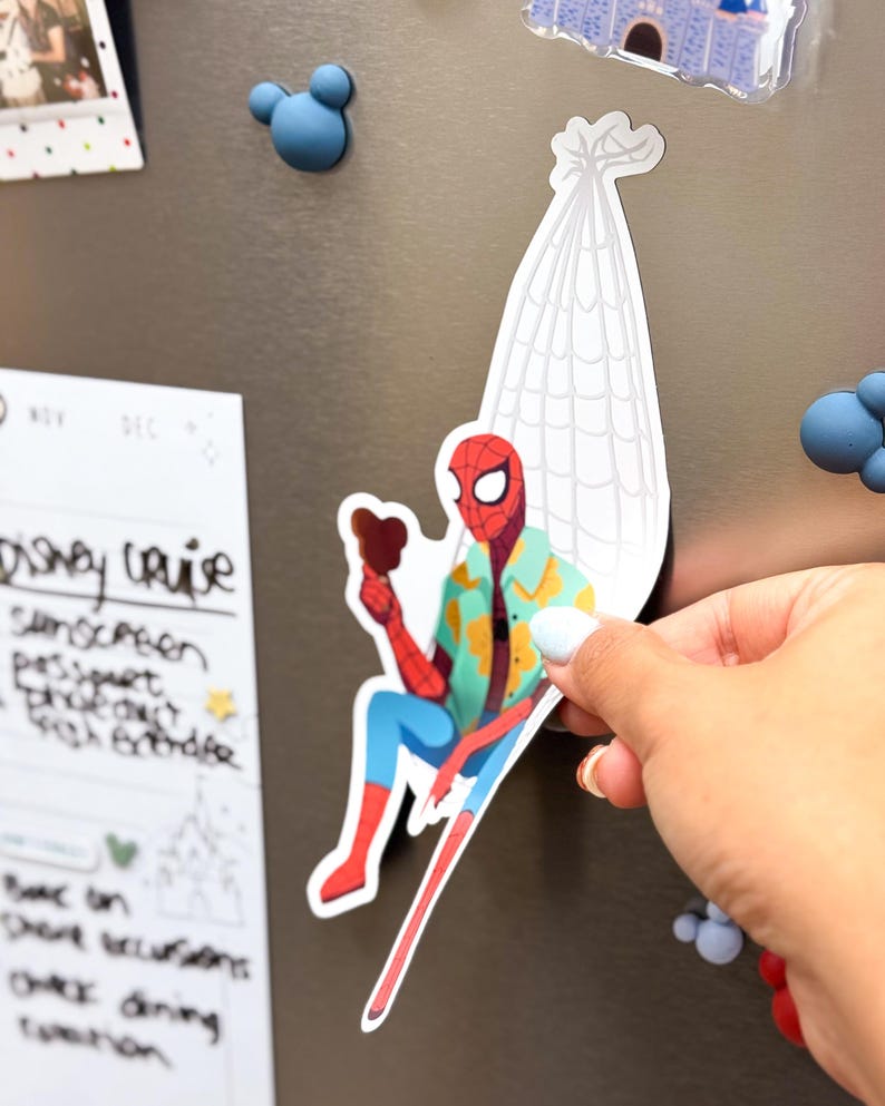 May include: A colorful Spider-Man magnet, holding a treat, is depicted sitting on a white web. The superhero is wearing a Hawaiian shirt, blue pants, and red boots. The magnet is being held up against a metallic surface.