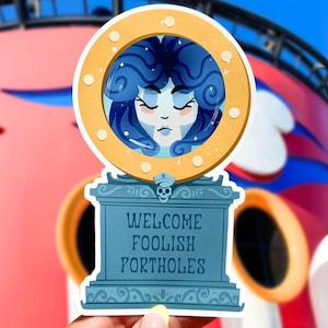 May include: A sticker featuring a stylized illustration of a woman with blue hair inside a gold-colored porthole design. The sticker has a blue base with the words "WELCOME FOOLISH PORTHOLES". The design includes a small skull and a sailor's hat.