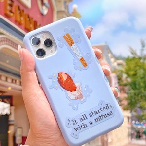 May include: A light blue phone case with decorative stickers of a churro, a wrapped pastry, and the text "It all started with a mouse." The case has a camera cutout and is held in a hand.