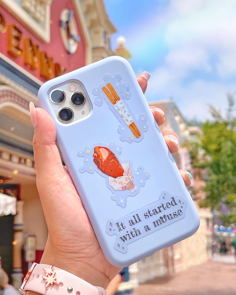 May include: A light blue phone case with decorative stickers of a churro, a wrapped pastry, and the text "It all started with a mouse." The case has a camera cutout and is held in a hand.