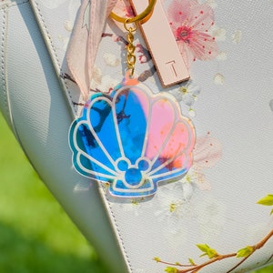 May include: A gold-tone keychain with a iridescent blue and pink seashell charm. The seashell has a Mickey Mouse silhouette inside.