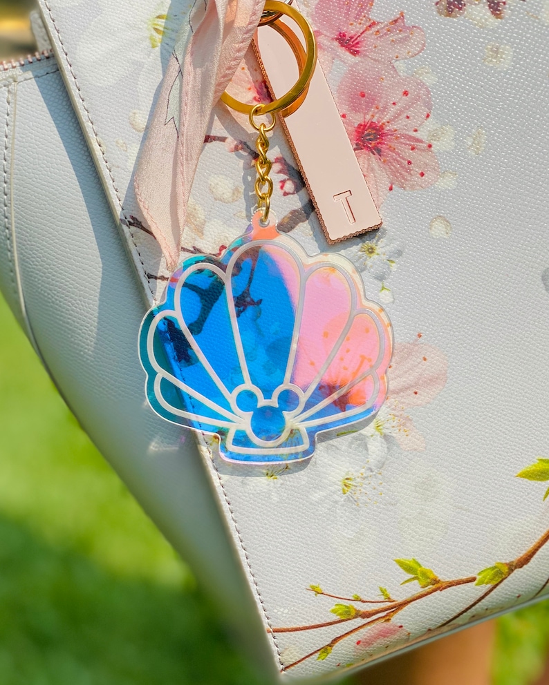 May include: A gold-tone keychain with a iridescent blue and pink seashell charm. The seashell has a Mickey Mouse silhouette inside.