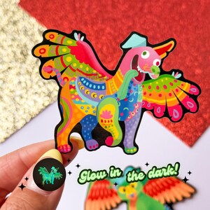 May include: A colorful, whimsical, glow-in-the-dark sticker of a mythical creature with wings. The creature is a mix of orange, yellow, pink, green, and blue colors. The sticker has a black outline and the text "Glow in the dark!" is written below it.