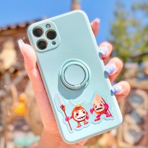 Pu&ograve; includere: Una custodia per telefono blu chiaro con un pop socket e un adesivo con due personaggi dei cartoni animati. I personaggi indossano abiti rossi e bianchi e hanno espressioni arrabbiate. La custodia &egrave; tenuta da una mano con smalto per unghie blu e bianco.