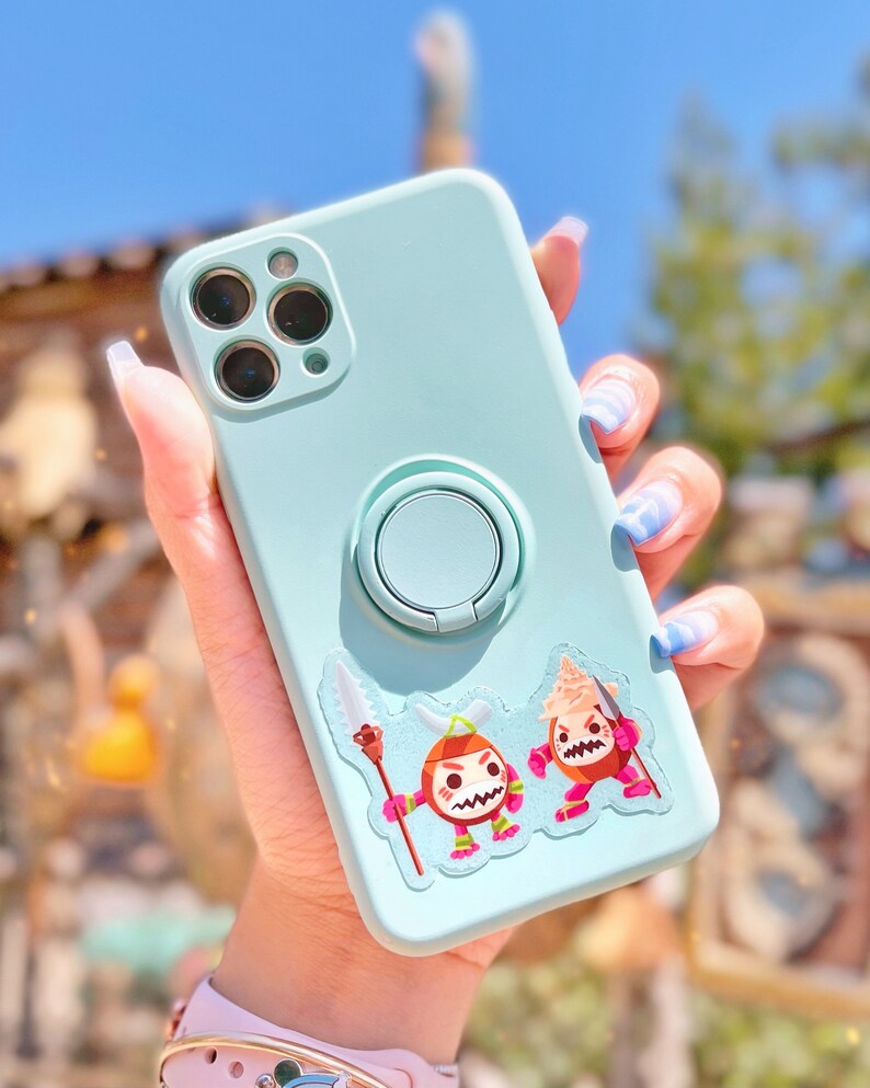 Pu&ograve; includere: Una custodia per telefono blu chiaro con un pop socket e un adesivo con due personaggi dei cartoni animati. I personaggi indossano abiti rossi e bianchi e hanno espressioni arrabbiate. La custodia &egrave; tenuta da una mano con smalto per unghie blu e bianco.