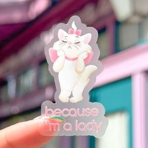 May include: A transparent sticker of a white cartoon cat with pink bows and a pink collar. The cat is holding its paws up to its face. The text "because I'm a lady" is printed in pink below the cat.