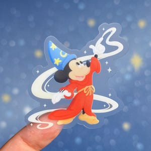 May include: A transparent sticker of Mickey Mouse dressed as a wizard, wearing a red robe and a blue hat with yellow stars. Mickey is conjuring a spell with a white swirl. The sticker is held against a blue backdrop.