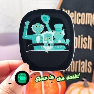 May include: A glow-in-the-dark sticker featuring three ghostly figures from the Haunted Mansion ride at Disneyland. The sticker is black with a green glow-in-the-dark effect. The text "Glow in the dark!" is written below the sticker.