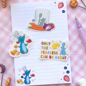 K&ouml;nnte beinhalten: Ein wei&szlig;es, liniertes Notizbuch mit bunten Aufklebern mit einer Cartoon-Kochratte, K&auml;se, Erdbeeren und Gem&uuml;se. Ein Aufkleber lautet "ONLY THE FEARLESS CAN BE GREAT". Ein lila Stift liegt daneben.