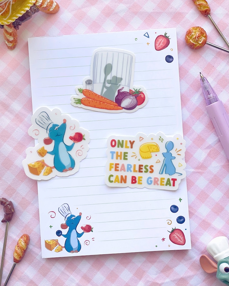 K&ouml;nnte beinhalten: Ein wei&szlig;es, liniertes Notizbuch mit bunten Aufklebern mit einer Cartoon-Kochratte, K&auml;se, Erdbeeren und Gem&uuml;se. Ein Aufkleber lautet "ONLY THE FEARLESS CAN BE GREAT". Ein lila Stift liegt daneben.