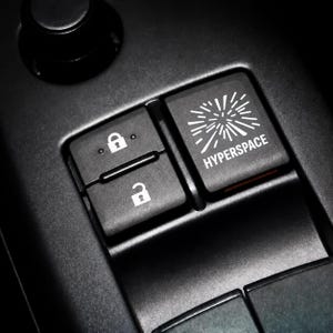 May include: A black car interior control panel featuring a rectangular button labeled HYPERSPACE with a white starburst graphic, positioned next to two smaller square buttons displaying white lock and unlock icons on a matte black surface.