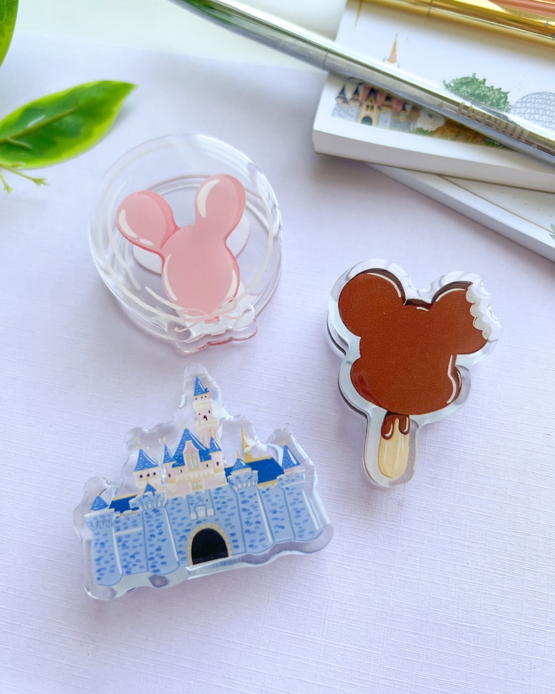 May include: Three clear acrylic pins with cartoon designs. One is a pink mouse head, another a brown ice cream bar, and the last a blue and white castle. The pins are displayed on a white surface.