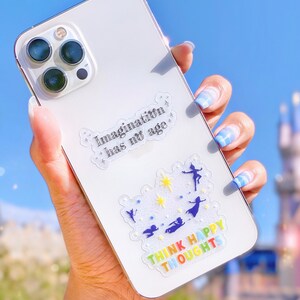 Op de afbeelding: Witte iPhone-hoes met een transparante sticker waarop "Imagination has no age" staat en een andere transparante sticker met een regenboogontwerp waarop "Think Happy Thoughts" staat.
