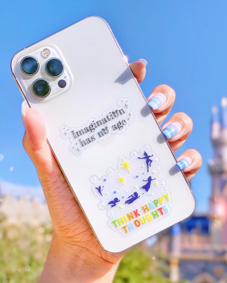 Op de afbeelding: Witte iPhone-hoes met een transparante sticker waarop "Imagination has no age" staat en een andere transparante sticker met een regenboogontwerp waarop "Think Happy Thoughts" staat.