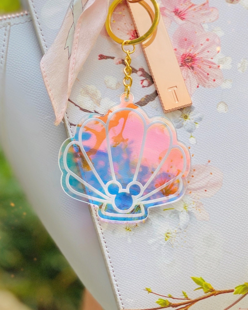 May include: A iridescent seashell-shaped keychain with a Mickey Mouse silhouette inside. The keychain is attached to a gold chain and a gold key ring.