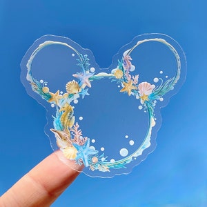 May include: Clear sticker with a Mickey Mouse shaped outline. The outline is decorated with seashells, starfish, and seahorses in shades of blue, yellow, and pink.