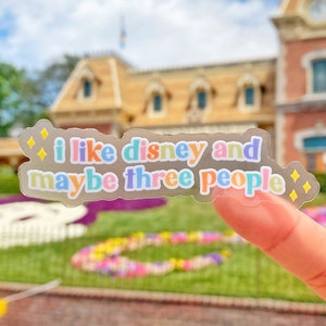 May include: A clear sticker with the text "i like disney and maybe three people" in rainbow colors with yellow glitter accents. The sticker is held up in front of a blurred background of a building with a brown roof.