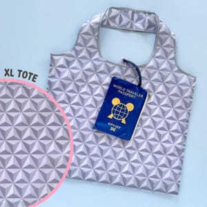 May include: An XL tote bag with a geometric pattern in shades of gray and white. A blue "World Traveler Passport" wallet with gold accents rests on the bag. The bag has a handle for carrying.