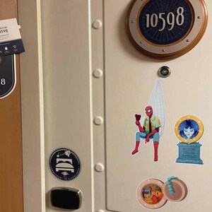 May include: A cream-colored door features a circular room number plaque with the number 10598. Decorative stickers include a Spider-Man figure, a welcome sign, and a nautical-themed design. A door handle and a sign are also visible.