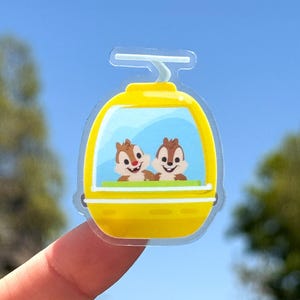 May include: A yellow and clear sticker featuring a gondola with Chip and Dale inside. The gondola has a blue window and a green seat. The background is a clear blue sky.