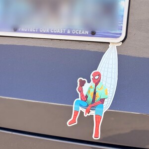 May include: A colorful sticker featuring Spider-Man relaxing in a hammock, holding a chocolate ice cream bar. He wears a Hawaiian shirt, blue pants, and red boots. The sticker is attached to a car near a license plate.