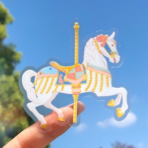 May include: A white carousel horse with a blue, pink, and gold saddle and a gold pole. The horse is standing on a clear background.