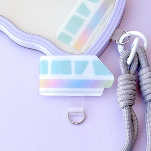 May include: A clear, iridescent train-shaped charm with a silver D-ring and a light blue and rainbow gradient design. The charm is attached to a lavender phone case and a gray lanyard. The background is a solid lavender color.