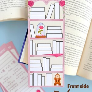 May include: A pink bookmark featuring a bookshelf design with white books, a rose in a glass dome, and a clock. The bookmark is decorated with pink roses at the top and is placed between the pages of an open book.