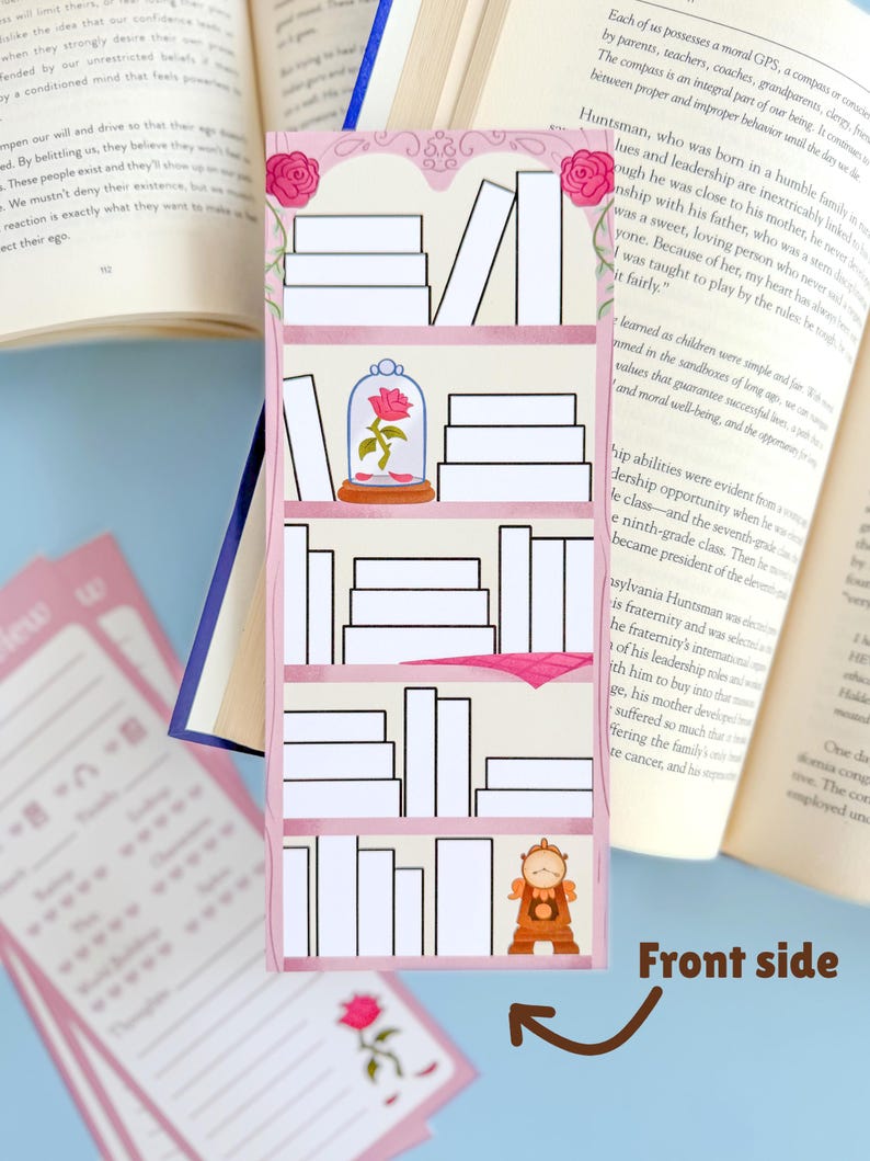 May include: A pink bookmark featuring a bookshelf design with white books, a rose in a glass dome, and a clock. The bookmark is decorated with pink roses at the top and is placed between the pages of an open book.