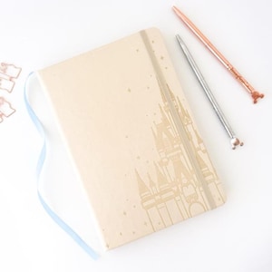 May include: A beige notebook with a castle design and a light blue ribbon. Two pens, one silver and one rose gold, are next to the notebook. The rose gold pen has a mouse-shaped end. A few rose gold paper clips are also visible.